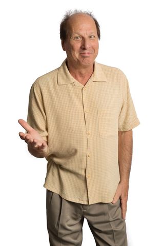 Adrian Belew wearing a yellow shirt and shrugging against a white background