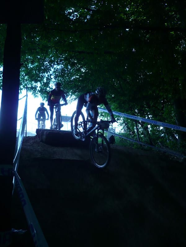 Racers come out of the sunlight and into the dark woods as they launch off this rock and drop in to some singletrack