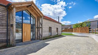 A striking barn-style home with timber cladding, a full-height glass entrance with a wooden front door, and a paved modern driveway under a bright blue sky.