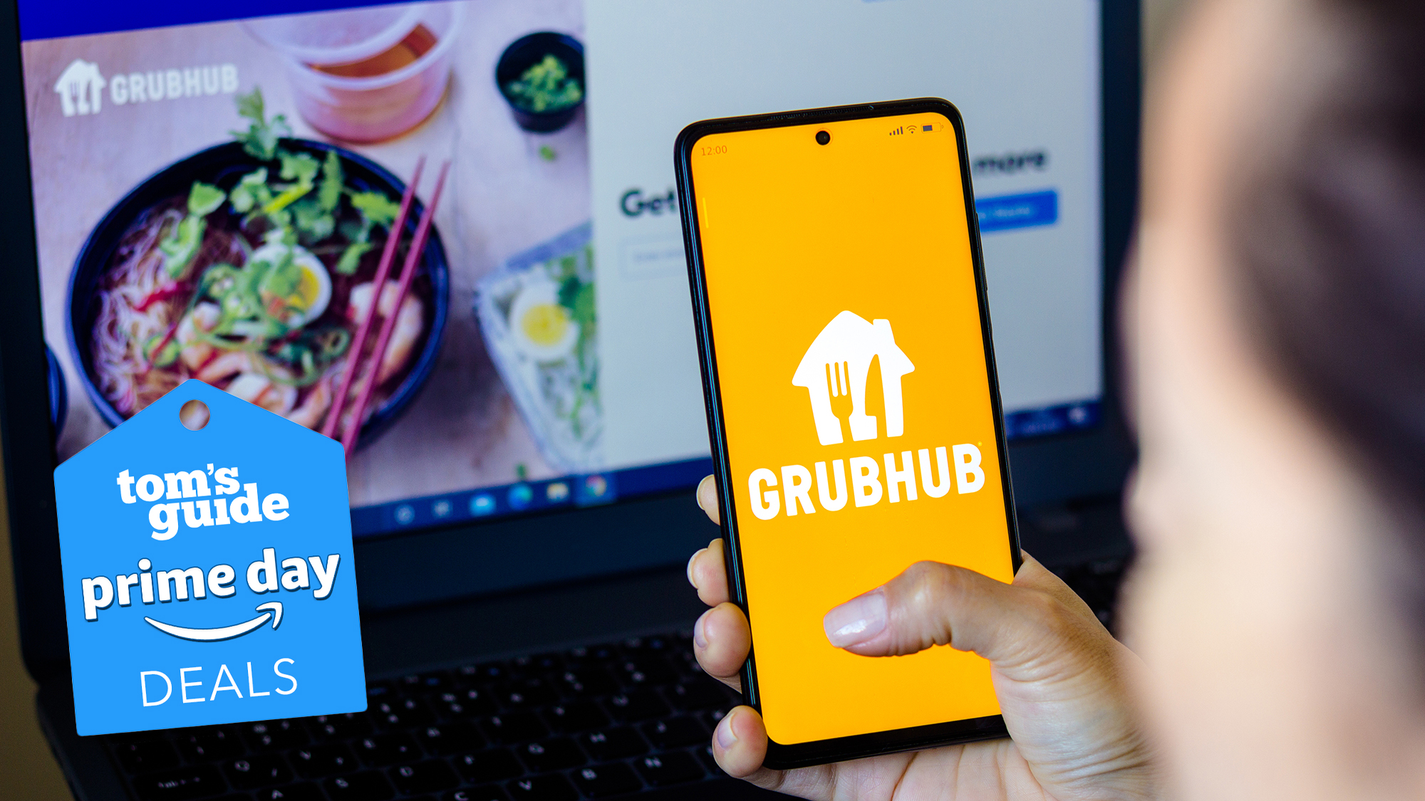 A phone with a Grubhub logo is in hand above a laptop with the Grubhub site