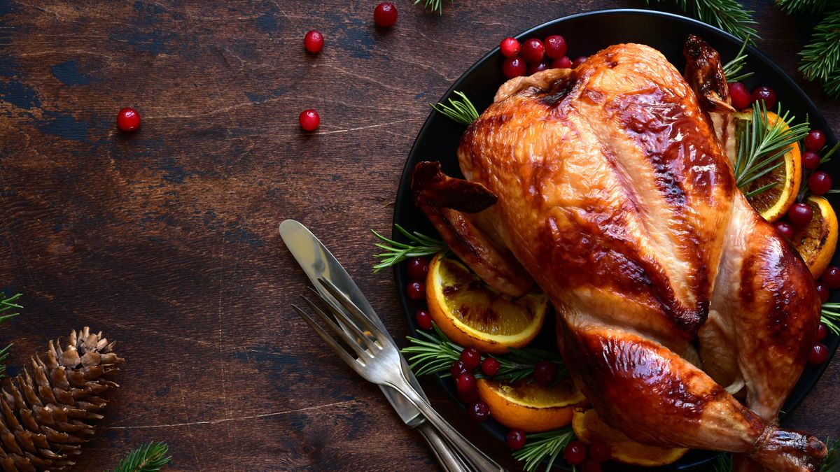 A cooked turkey on a plate with oranges, cranberries and thyme, next to a knife and fork