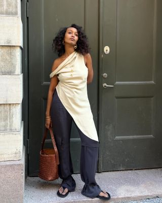 Gaya Musim Panas yang Elegan: @monamali_ mengenakan atasan krem ​​​​asimetris dengan celana panjang navy dan tas kulit tenun berwarna coklat