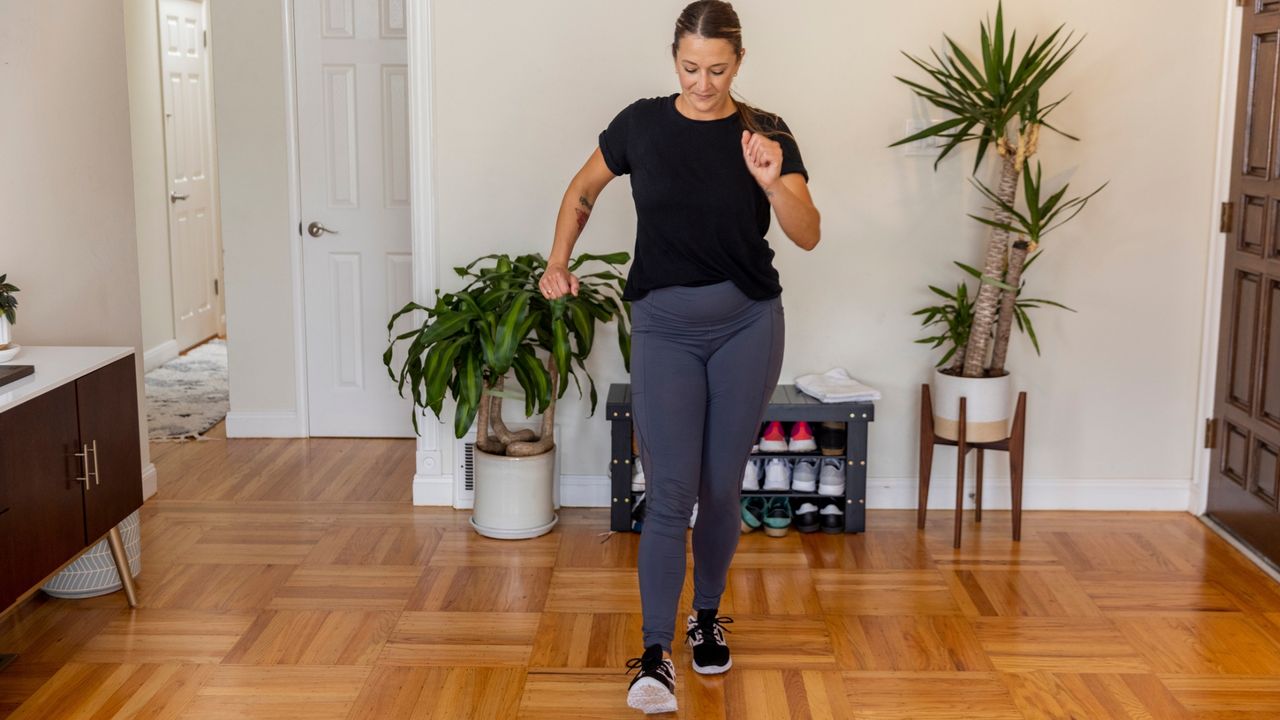 white woman exercising at home doing standing heel tap movement, on wooden floor and plants in the background