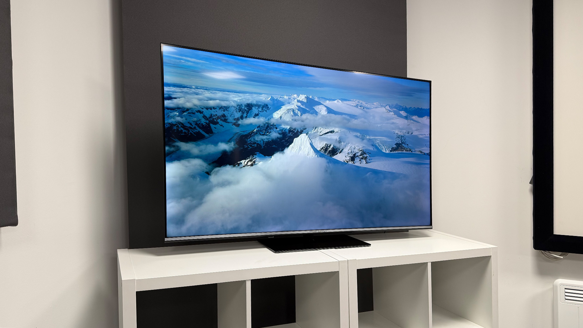 Mitchell &amp;amp; Brown QLED1811 QLED TV on white shelving unit, on screen is aerial view of mountains and clouds