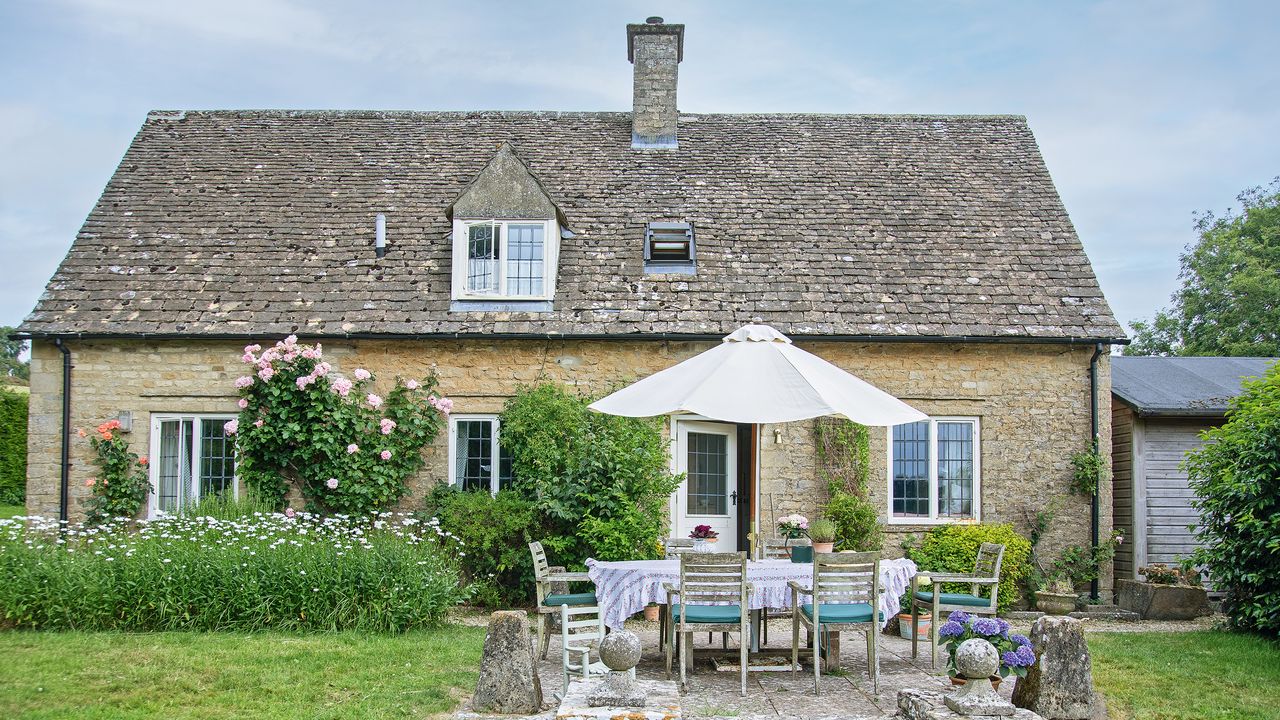 exterior of a stone cottage with roses up the walls and terrace area with parasol and table