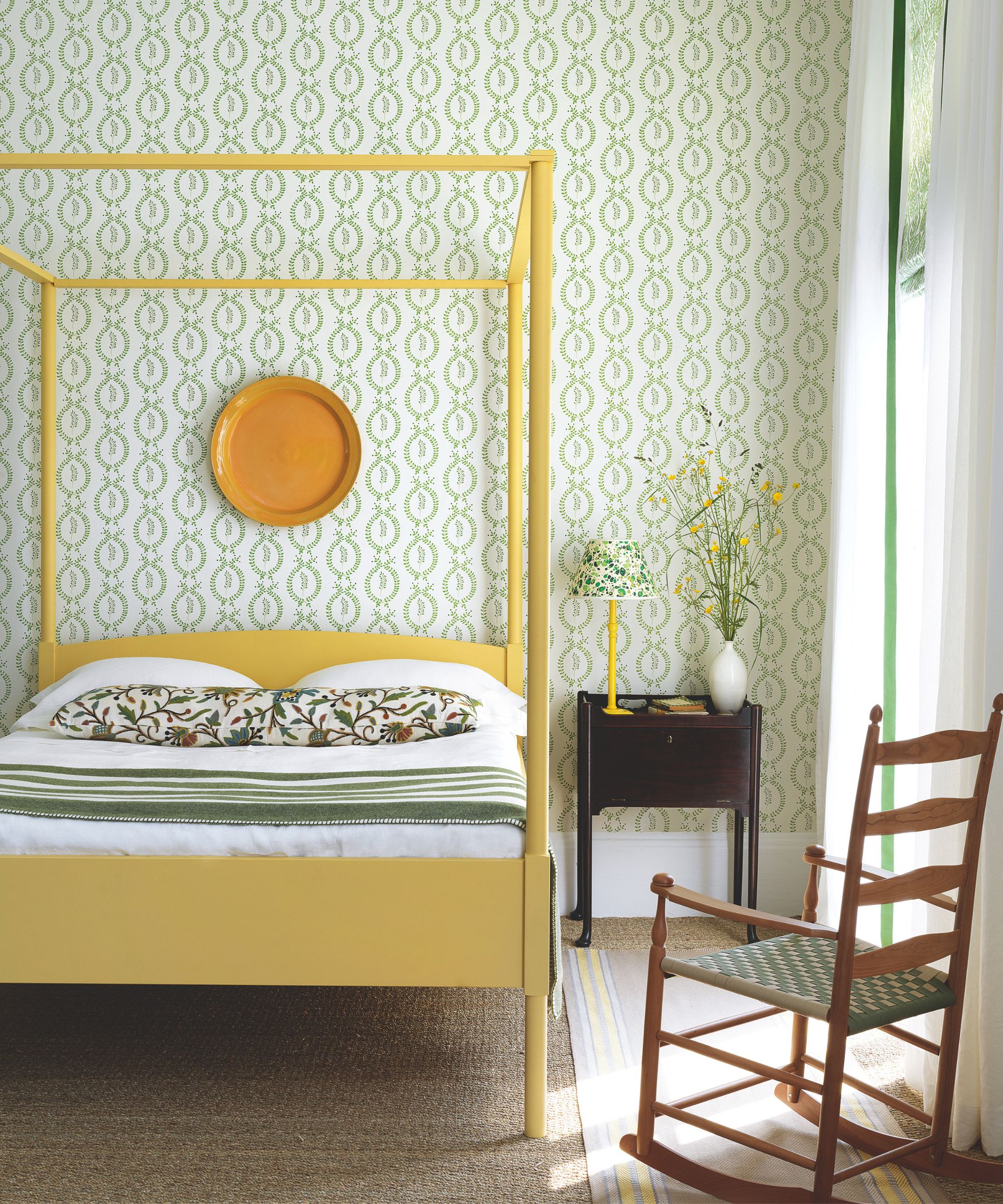 Bright bedroom with green wallpaper and yellow four poster bed, with white bedding, a long floral bolster cushion, and a green and white striped blanket, brown nightstand and wooden rocking chair.