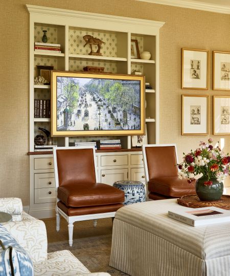 Living room with white built-in bookcase housing books and decor, with a TV mounted in front disguised as a painting, two brown leather chairs in front with a striped fabric ottoman at the center decorated with a rattan tray and a vase of flowers