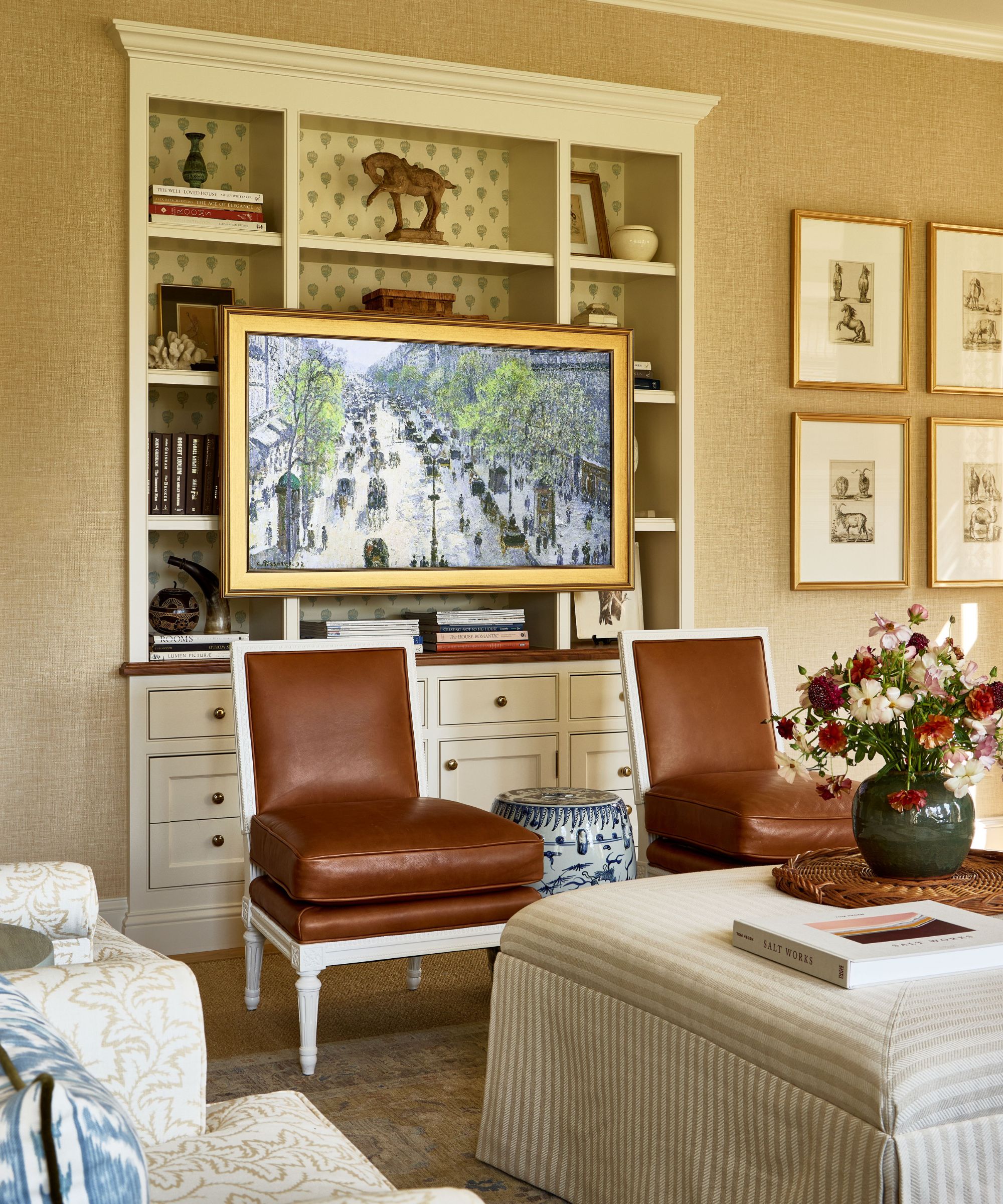 Living room with white built-in bookcase housing books and decor, with a TV mounted in front disguised as a painting, two brown leather chairs in front with a striped fabric ottoman at the center decorated with a rattan tray and a vase of flowers