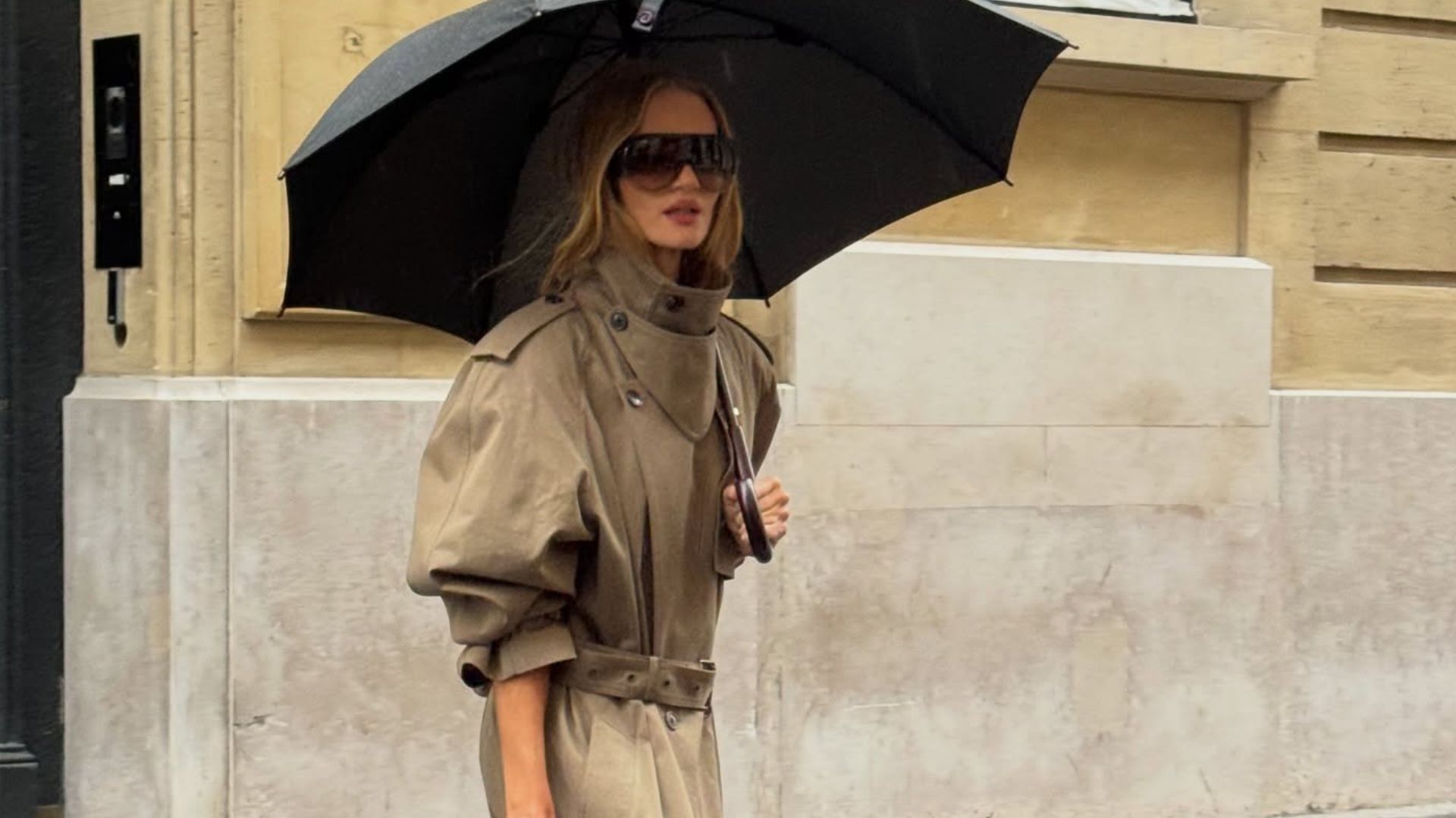 Model Rosie Huntington-Whiteley walks outside in the rain underneath an umbrella, wearing a beige funnel-neck trench coat with burgundy boots and a bowling bag.