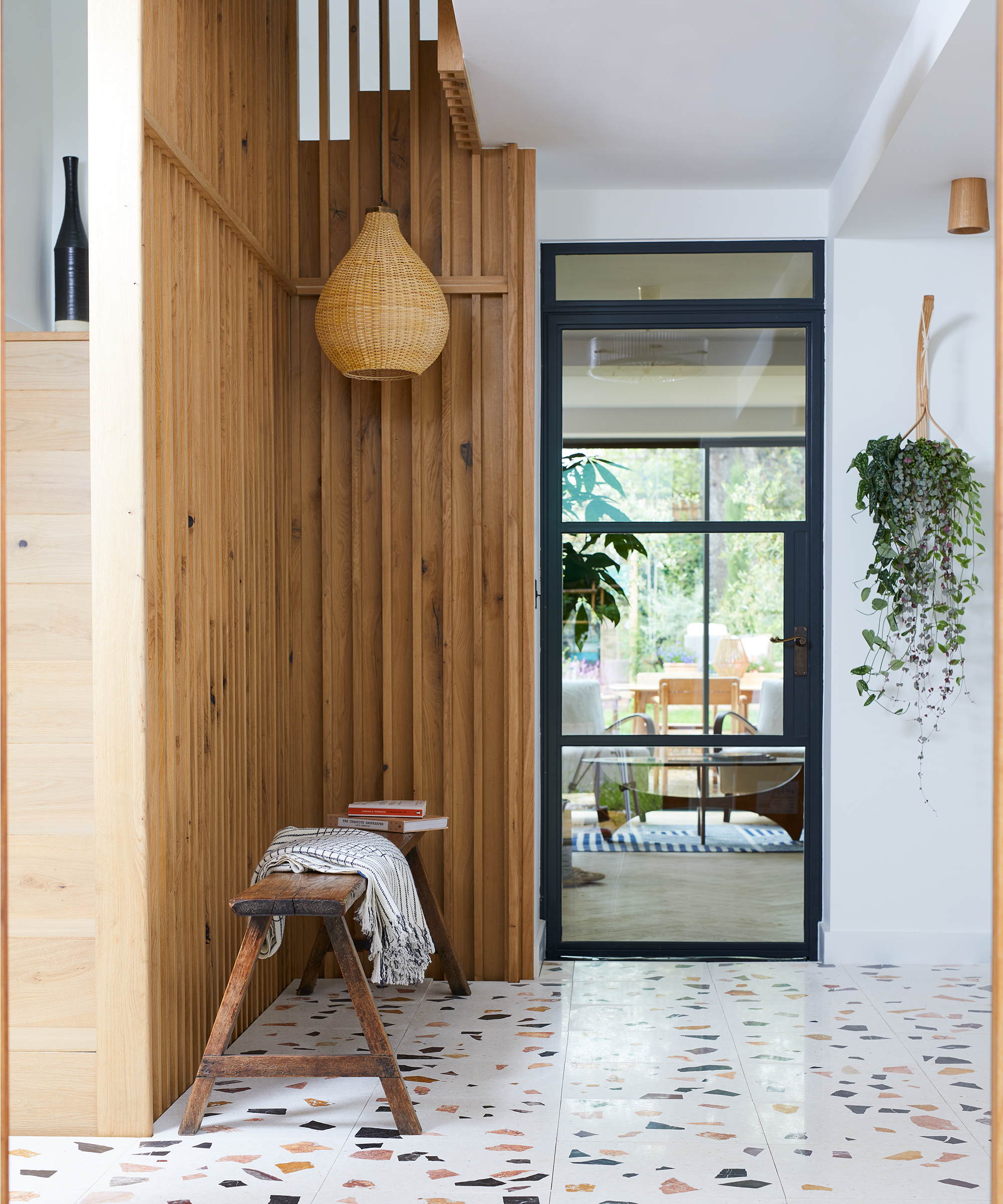 modern hallway with vertical wood panelling feature on wall, hanging rattan pendant light, small bench, terrazzo style floor tiles and black framed glazed door leading into another room