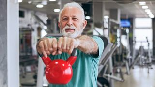 A man in a gym performs a kettlebell front raise. He's facing the camera and photographed from the waist up. His arm are stretched out straight in front of him, with his hands clasping the handle of the red kettlebell.