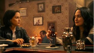 Rebecca Godfrey (Riley Keough) and Officer Cam Bentland (Lily Gladstone) look inquiringly at one another as they sit nursing their drinks in a warmly lit bar in Victoria, British Columbia, in true crime series Under the Bridge.