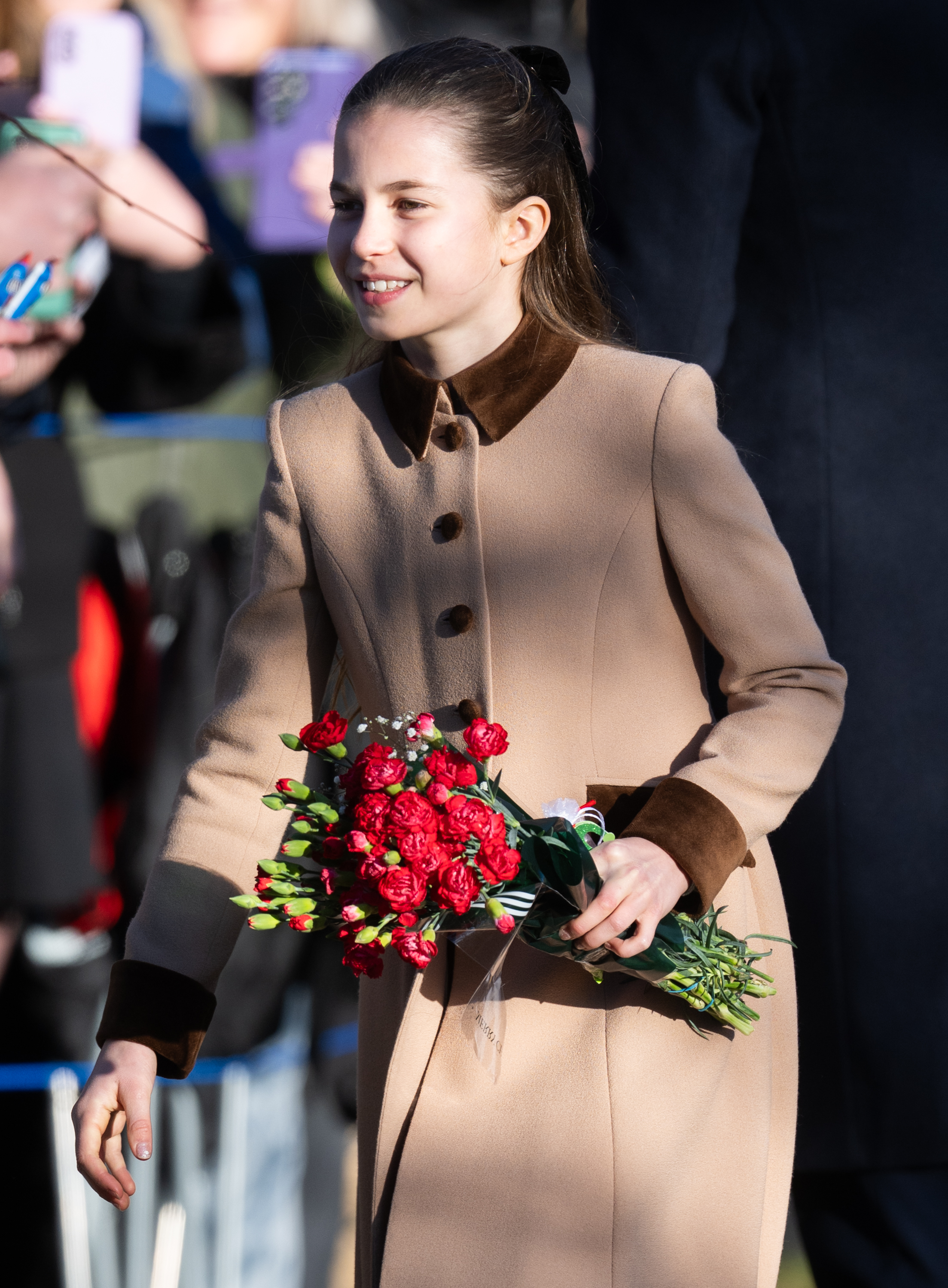 Princess Charlotte wears a light brown coat on Christmas Day in 2025 and is handed yellow and red flowers by royal fans in Sandringham