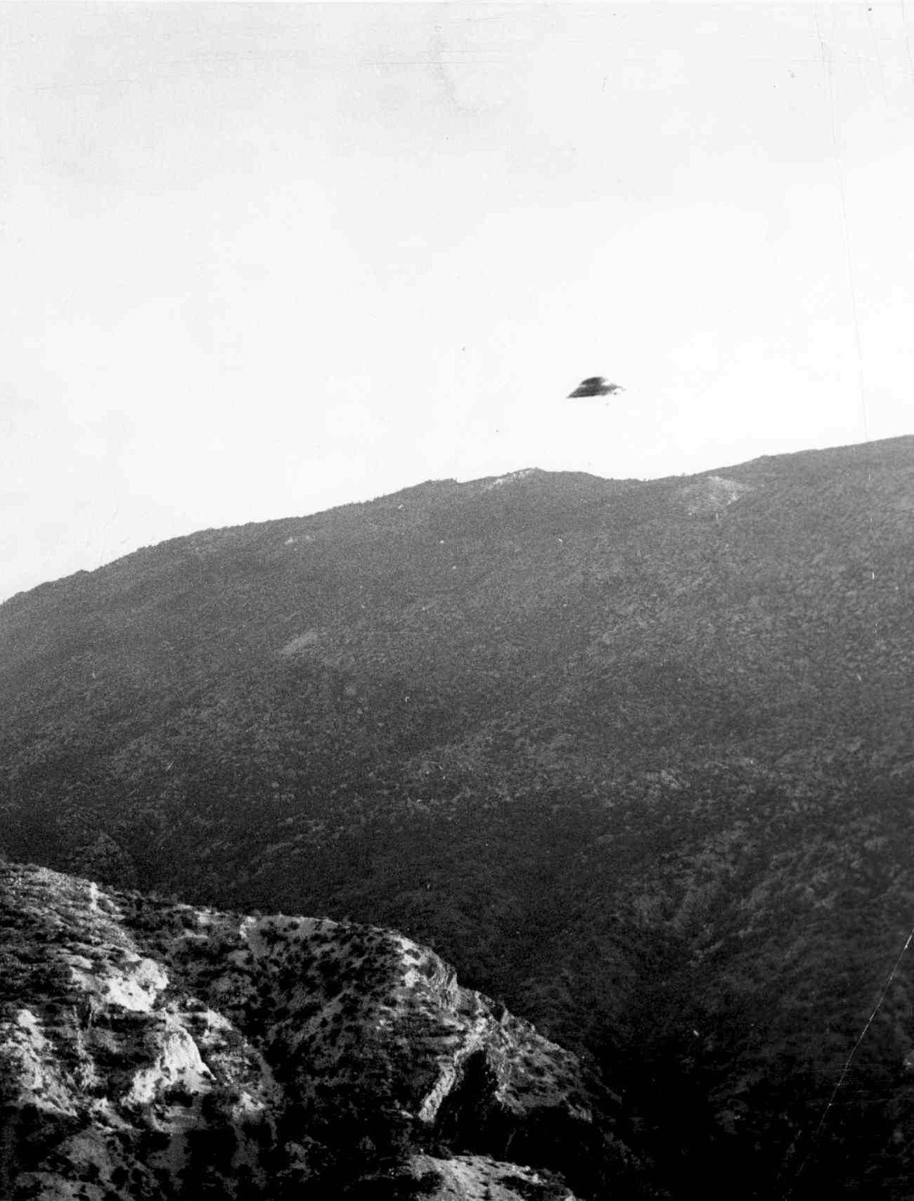 a ufo flies through the sky above a large forested hill in black and white.