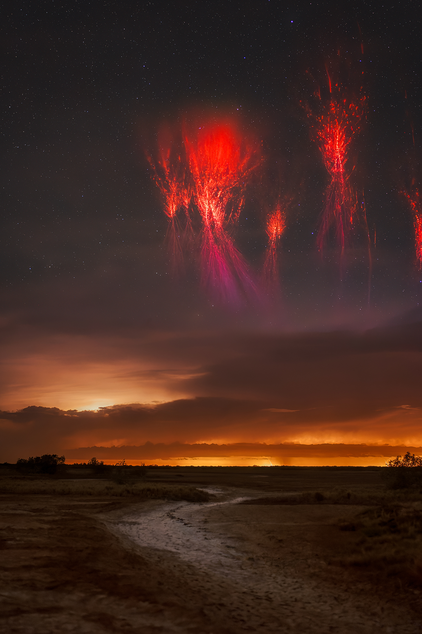 A vibrant night sky displays red lightning-like sprites above a dark landscape, contrasted by a subtle orange horizon. Stars twinkle overhead
