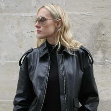 Woman wearing black turtleneck, black leather jacket, and sunglasses standing against Parisian wall.