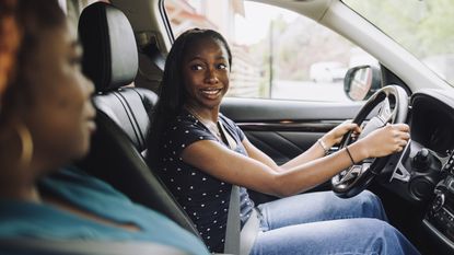 A teenager in the driver seat of a car looks over at her mother in the passenger seat.