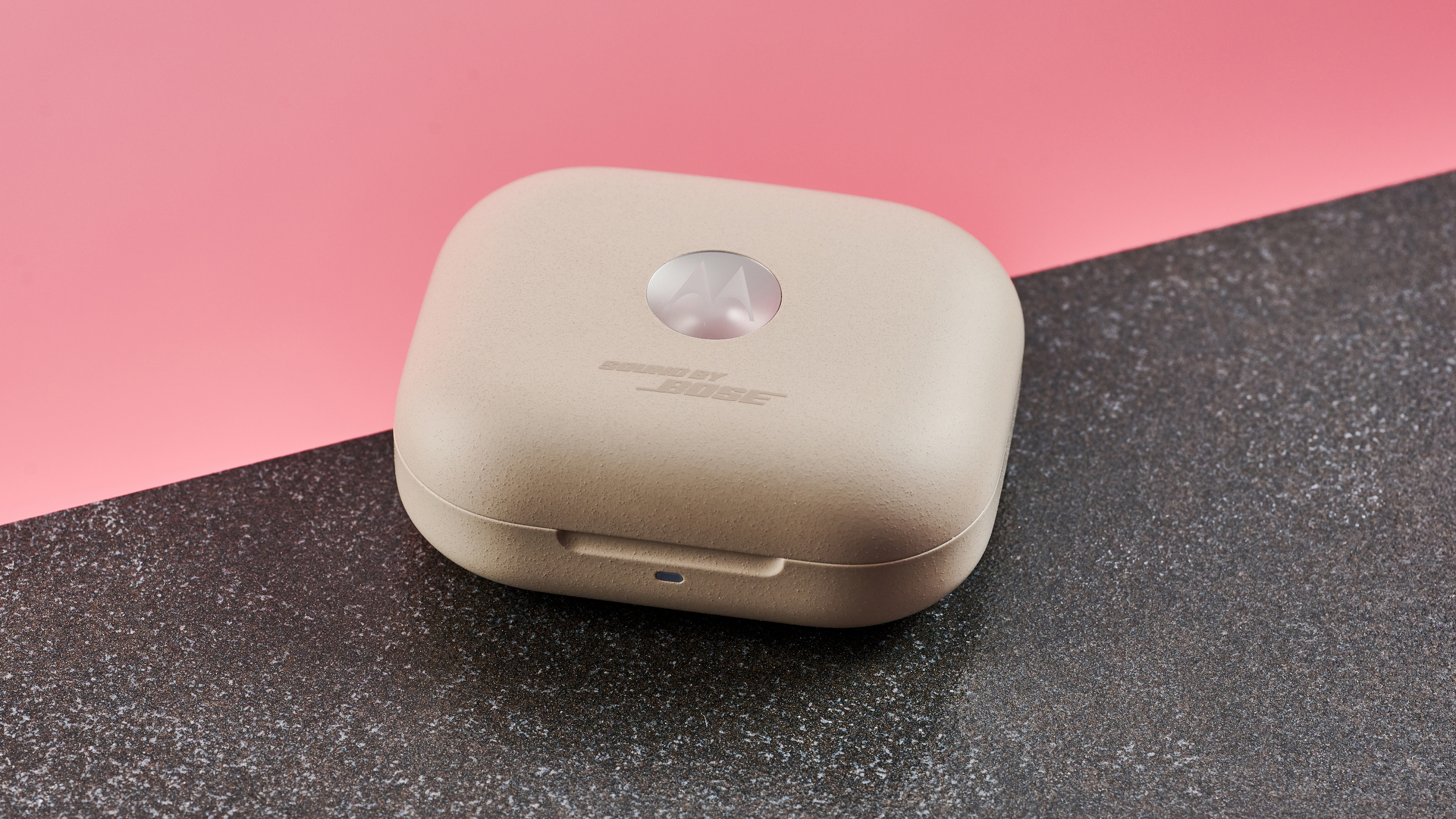 The wireless Motorola Moto Buds Plus case is closed, showing the silver Motorola logo on the top. The case is a sandy colour and has a rough texture. It is pictured on a dark surface and against a pink background.