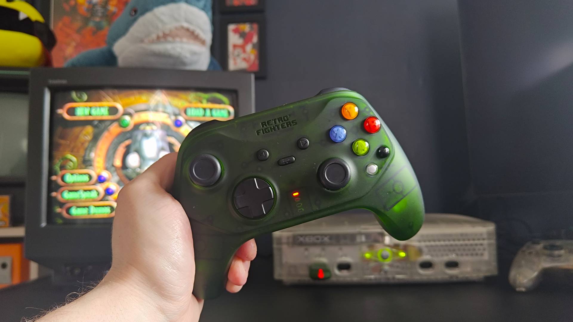 Hand holding Retro Fighters Hunter with OG Xbox Crystal Edition and Sony Trinitron TV in backdrop with Oddword: Munch's Odyssee main menu on screen.