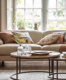 A spring themes living room with a large cream fabric sofa behind two round coffee tables. A glass vase of yellow flowers on the table. Assorted throw cushions on the sofa. A curved wooden floor lamp visible in the right hand corner. 