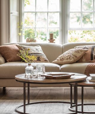 A spring themes living room with a large cream fabric sofa behind two round coffee tables. A glass vase of yellow flowers on the table. Assorted throw cushions on the sofa. A curved wooden floor lamp visible in the right hand corner.