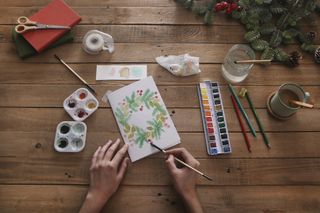 A person painting a Christmas wreath in watercolours onto a white piece of card, surrounded by art supplies such as watercolour paints and paint brushes.