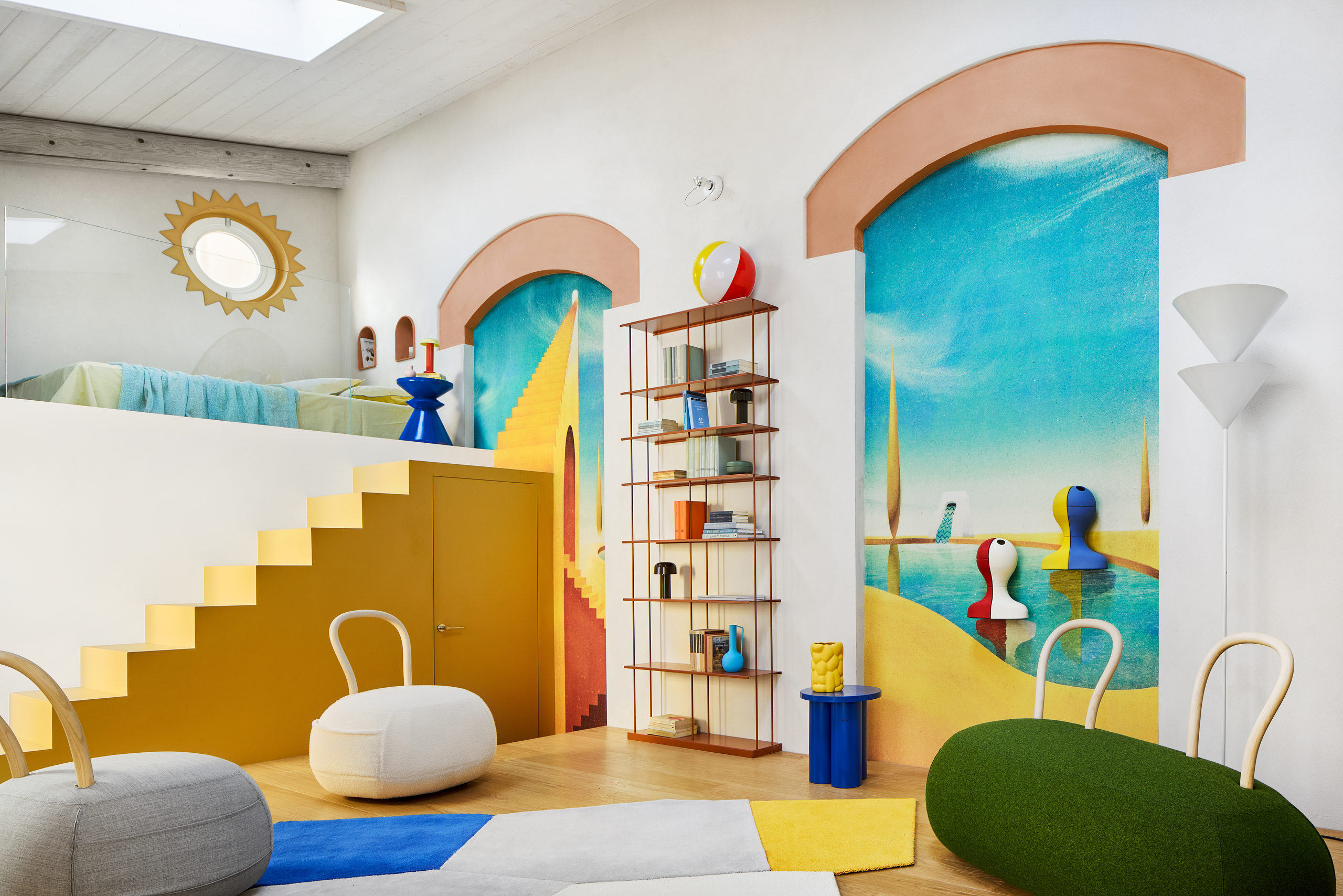A white studio apartment with a colorful mural, arched moulding, an open shelf of colorful decor, a set of yellow stairs leading to a loft bed, and Yum Yum chairs by Opinion Ciatti