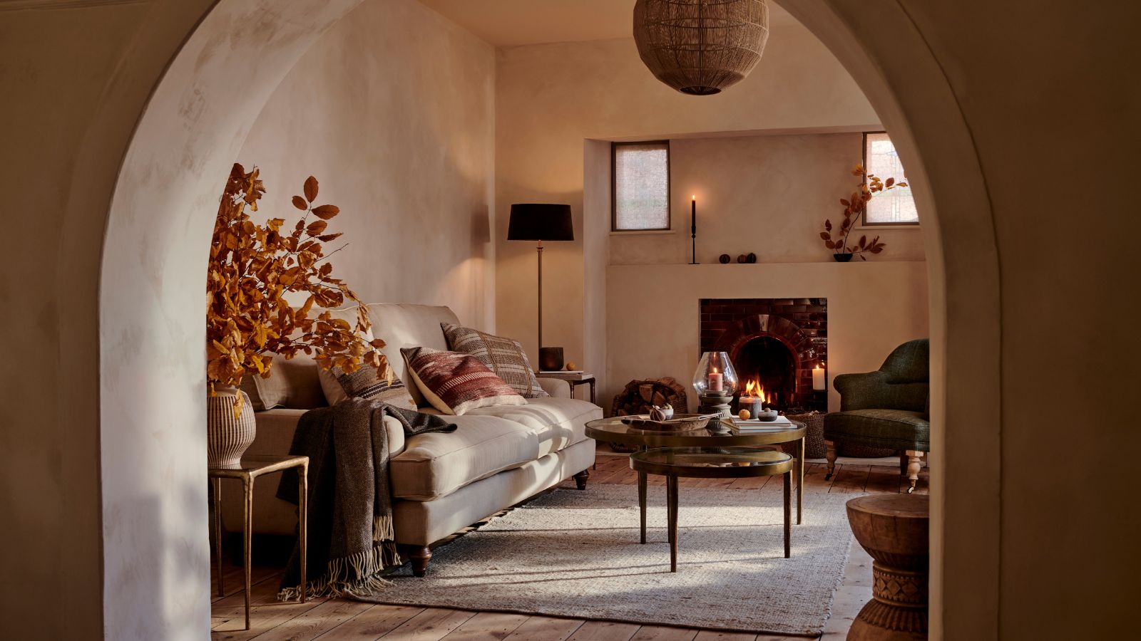 A cozy fall-esque living room viewed through an archway. A cozy cream sofa with green and red throw cushions behind a round coffee table. A large orange foliage arrangement in a vase beside it. 