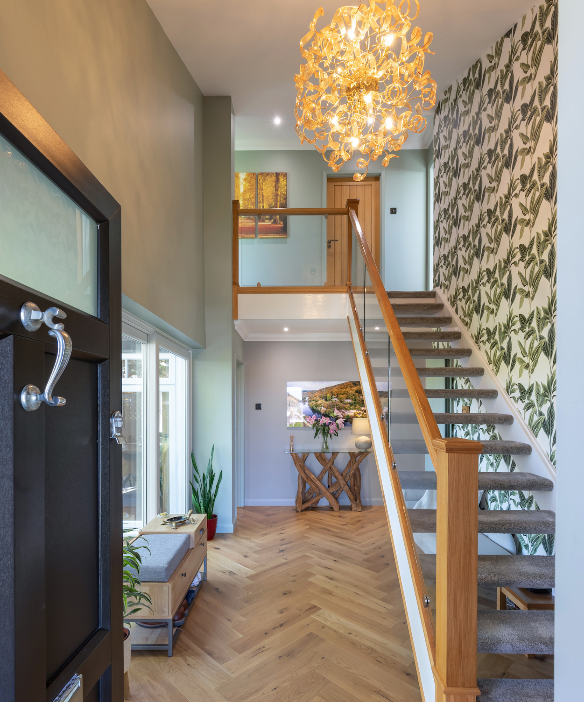 large hallway with open tread staircase, glass balustrade, wooden floor, floral patterned wallpaper, green wall and large modern pendant light