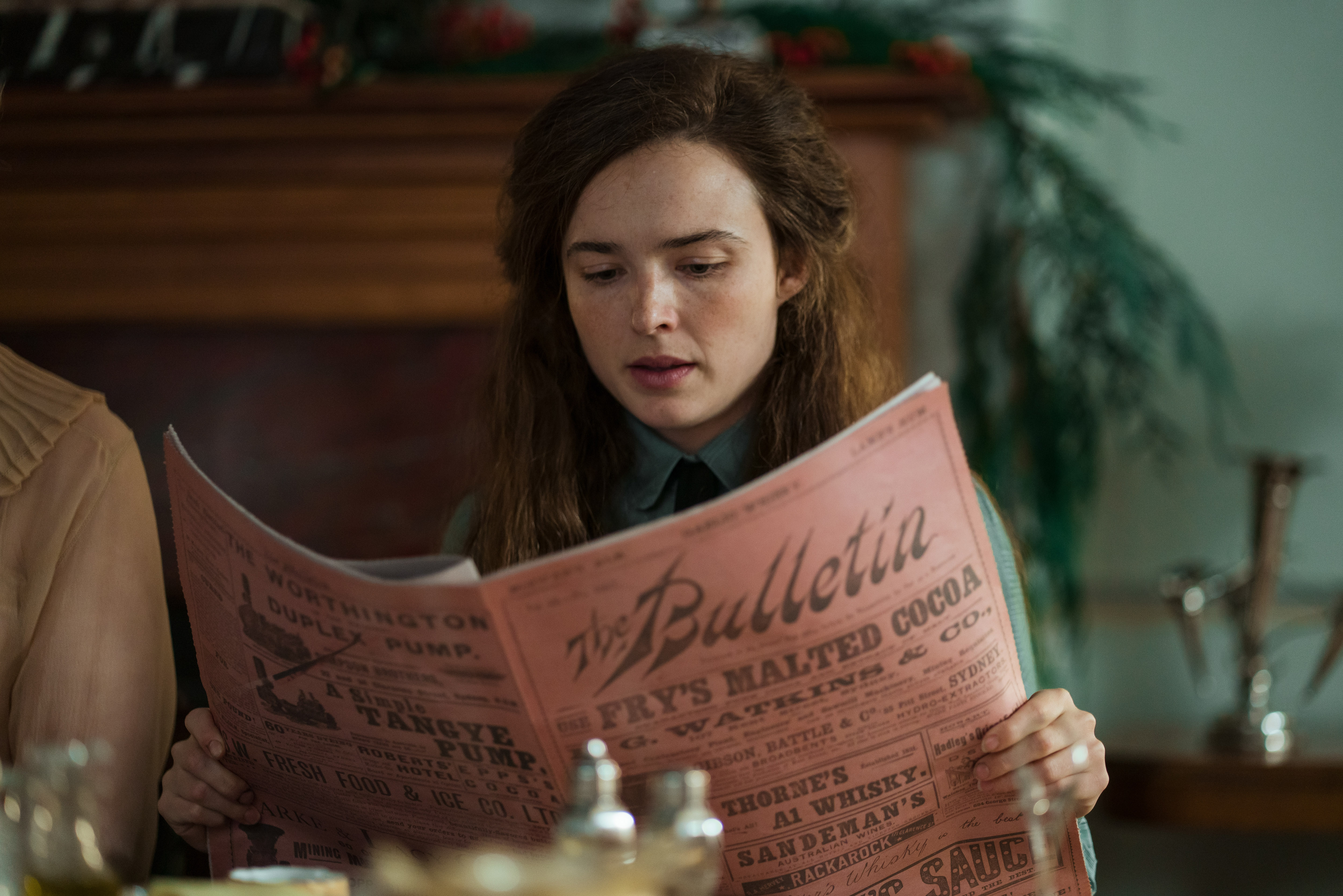 Phillipa Northeast as Sybylla, reading a 20th-century-era newspaper as she sits at a dining table, in the Netflix period drama 'My Brilliant Career.'