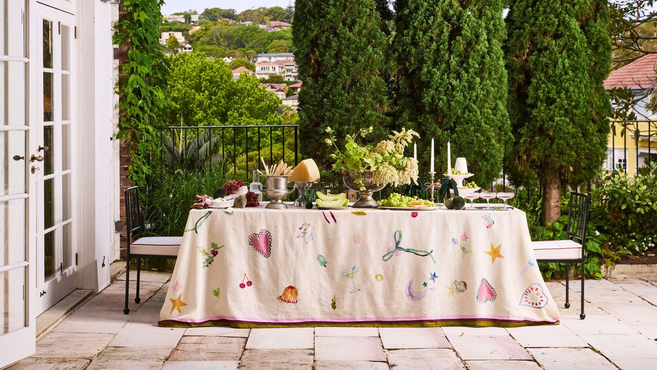 Bed Threads annie everingham collaboration tablecloth with drawn-on motifs on a table styled with silverware, plates, candles, and cheese platters, with views of trees in the background