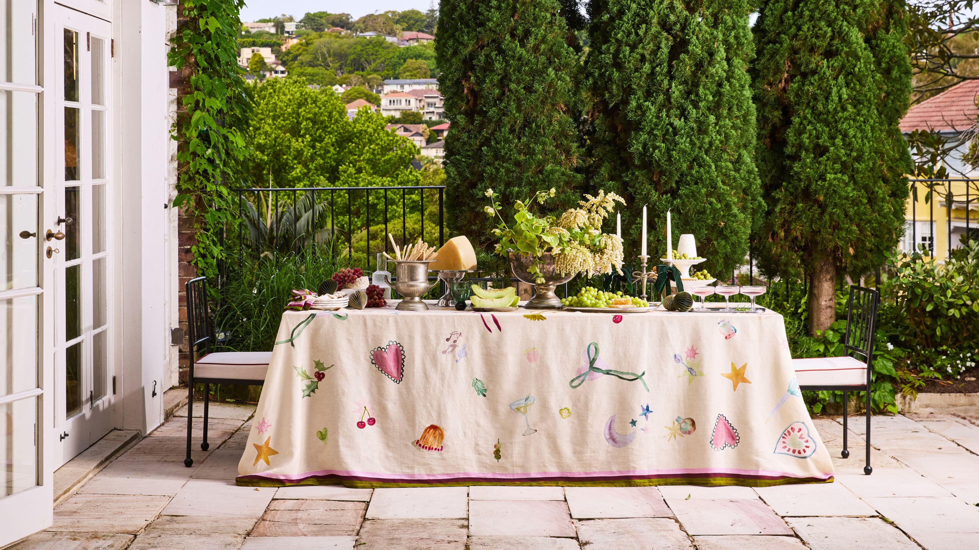 Bed Threads annie everingham collaboration tablecloth with drawn-on motifs on a table styled with silverware, plates, candles, and cheese platters, with views of trees in the background