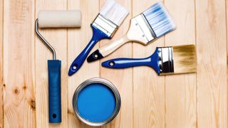 Top down shot of mini roller and three paintbrushes with blue handles and small opened tin of blue paint on pine wood background