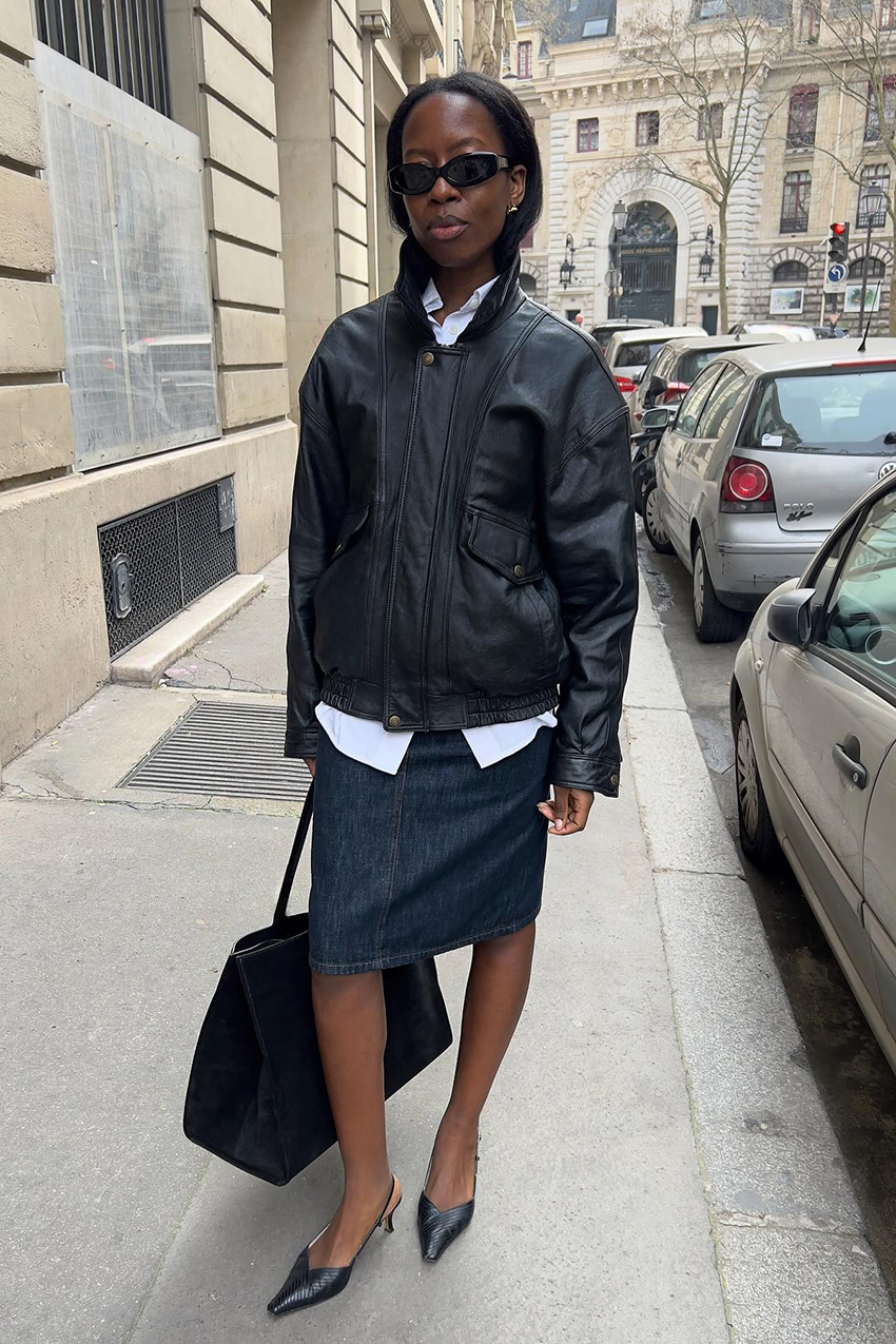 a woman wearing a leather jacket, denim skirt, white button-down shirt, and pointed toe heels
