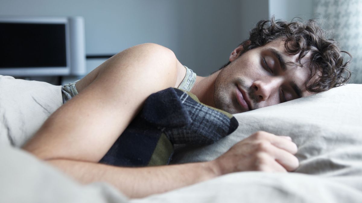 A man with short and curly brown hair sleeps peacefully on his side in bed