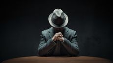 A man wearing a fedora sits with his head down so you can't see his face and his hands folded. 