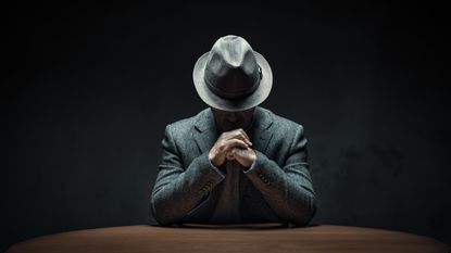 A man wearing a fedora sits with his head down so you can't see his face and his hands folded. 