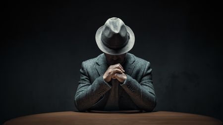 A man wearing a fedora sits with his head down so you can't see his face and his hands folded. 