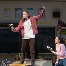 sadie sink stands on a table holding a book while another girl looks on in a shot of the play john proctor is the villain
