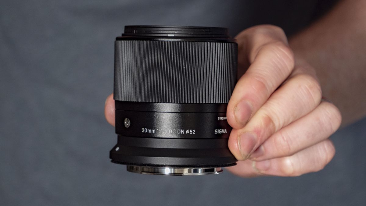 Sigma 30mm F1.4 DC DN Contemporary lens in the hand, blue t-shirt background