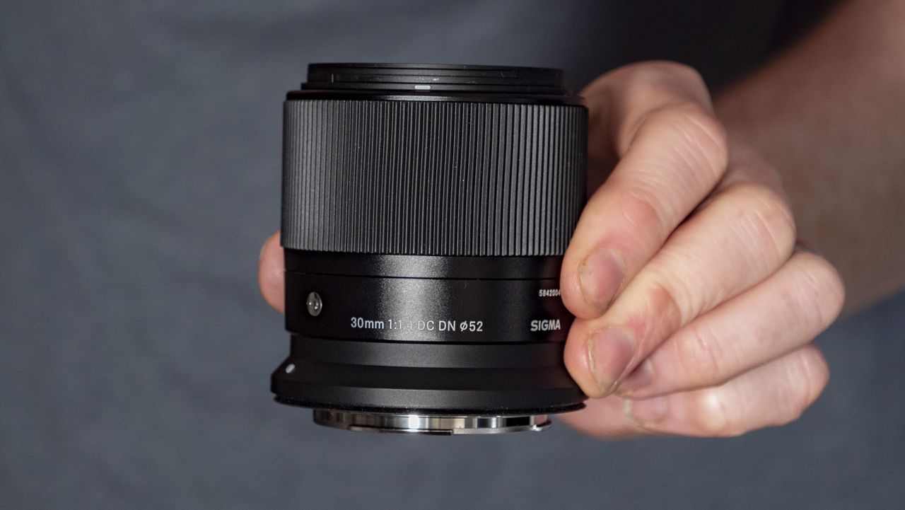 Sigma 30mm F1.4 DC DN Contemporary lens in the hand, blue t-shirt background