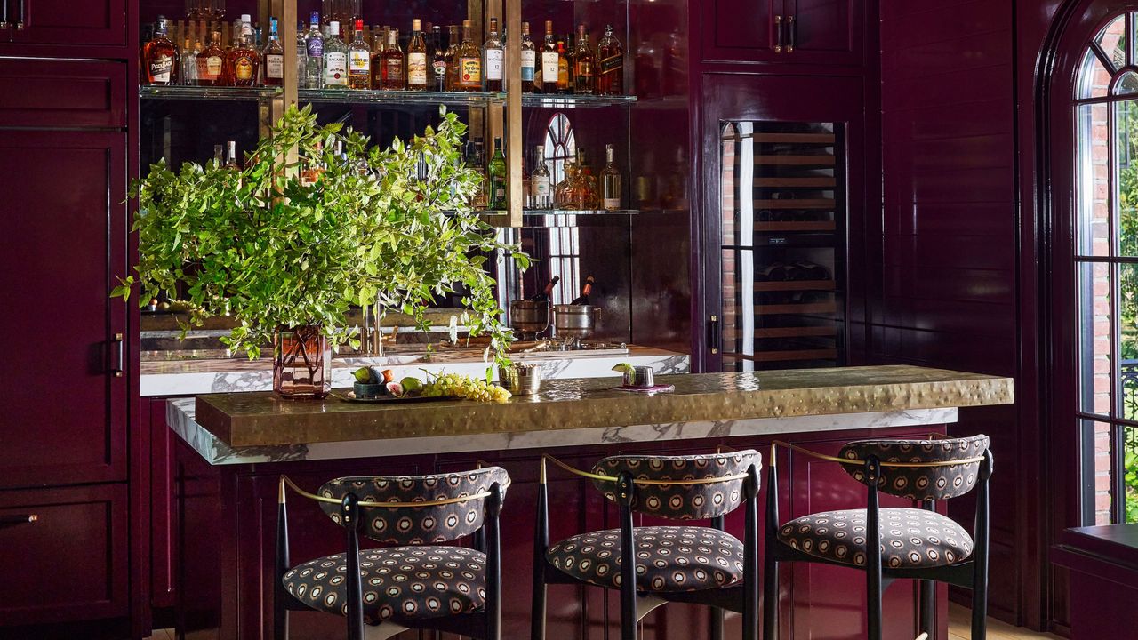 burgundy color drenched home bar with marble counter, three stools, a vase with green foliage and a mirrored splashback with shelves lined with spirits