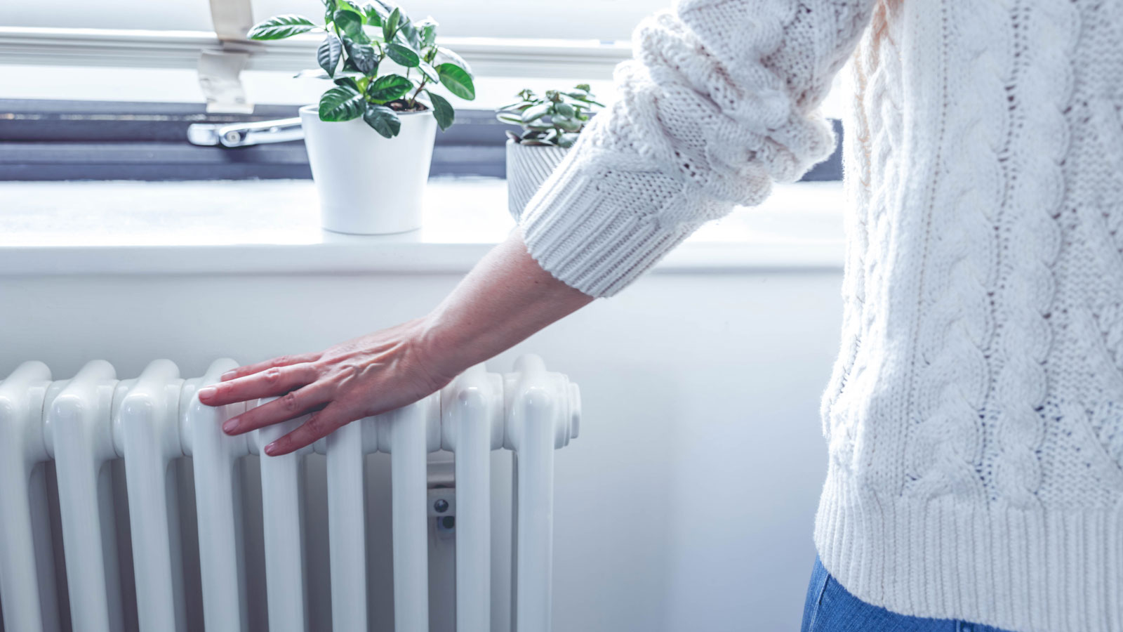 Person in white jumper and jeans running hand over white column radiator under window