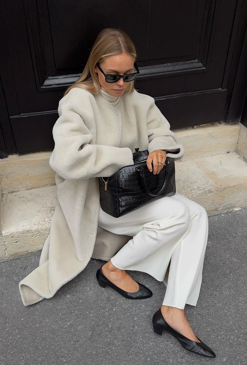 A woman sitting on a step wearing black sunglasses, a white shearling coat over a white turtleneck, white wide-leg trousers, black kitten heel pumps, and a black bowler bag.