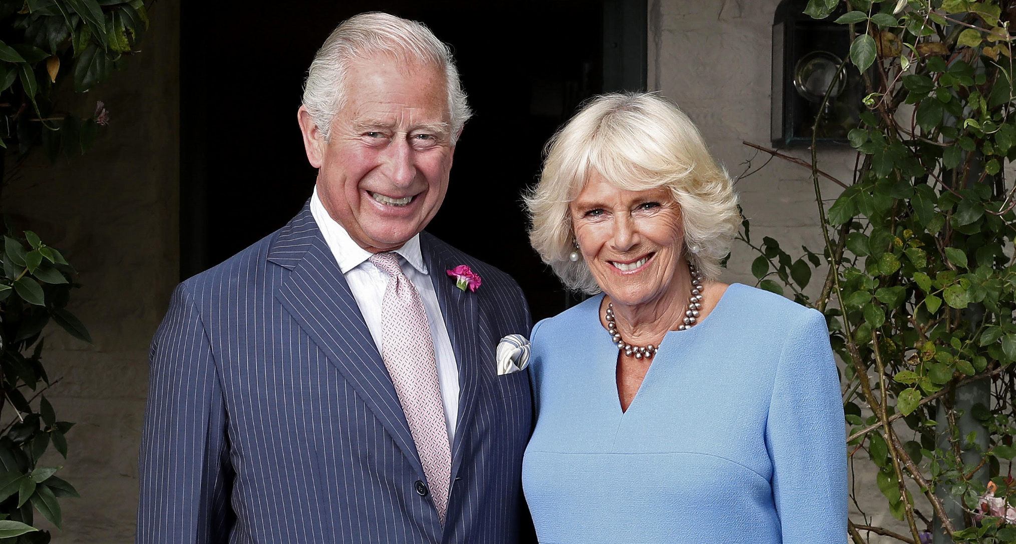 King Charles in a striped blue suit standing next to Queen Camilla, in a blue dress, and smiling 