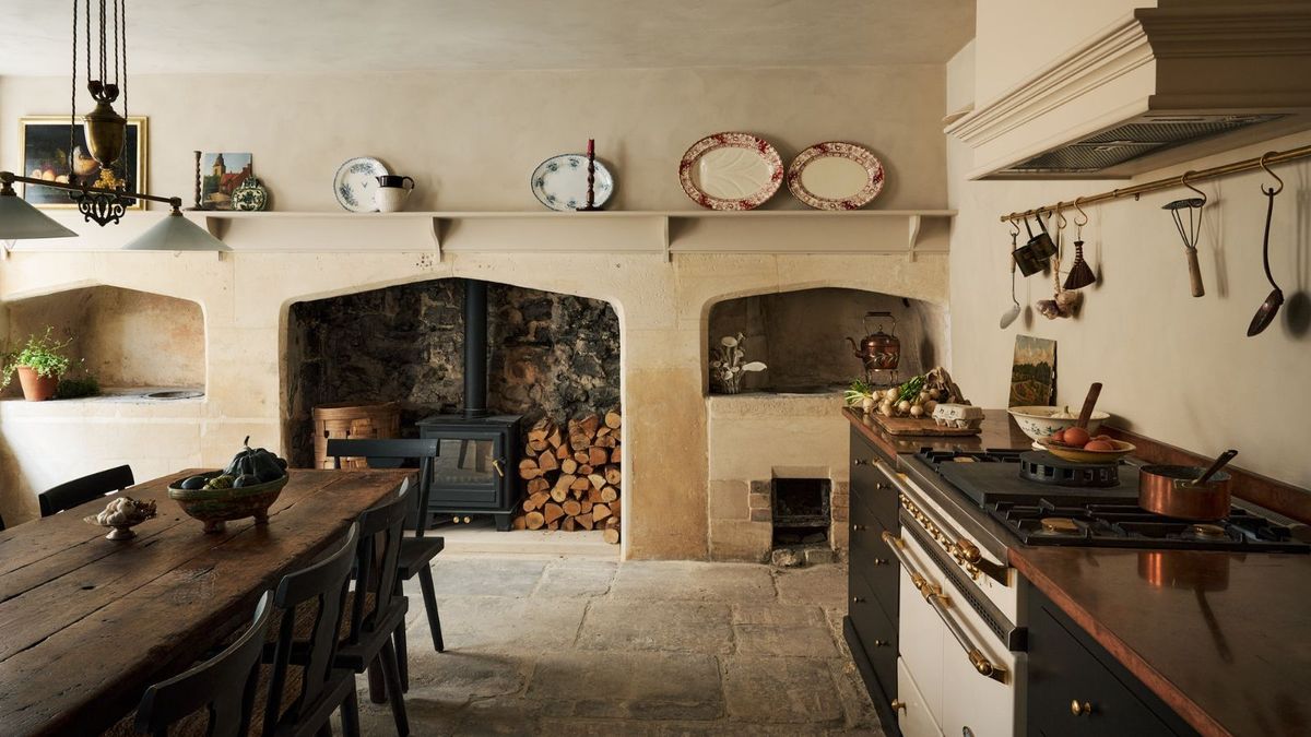 A rustic kitchen with a historic charm, featuring a large stone fireplace with log storage and a wood-burning stove. The kitchen includes a traditional range cooker with a copper worktop and dark cabinetry, adorned with brass hardware.