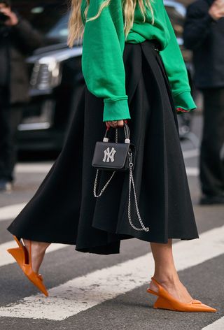 a street style photo of a woman wearing a green sweater with a black skirt and orange pumps and a mini phone bag