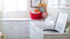 Open top-loading washing machine on right side of frame situated in a white kitchen with bright window and red cabinet pulls