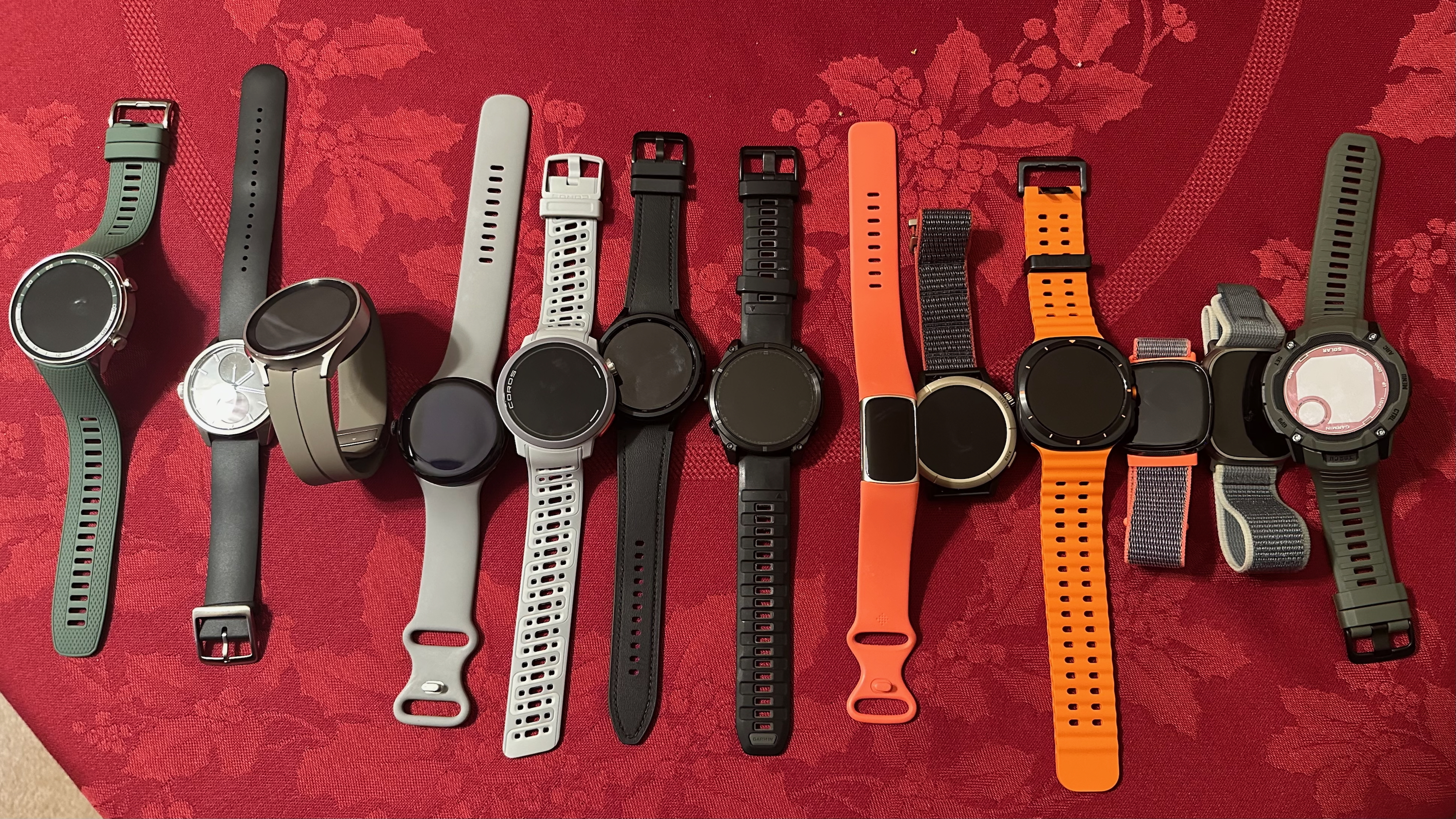 A top-down view of thirteen smartwatches sitting side-by-side on a desk. In order, they are the OnePlus Watch 2R, Withings ScanWatch 2, Samsung Galaxy Watch 5 Pro, Google Pixel Watch 3, Coros Pace Pro, Samsung Galaxy Watch 6 Classic, Garmin Fenix 8, Fitbit Charge 6, Amazfit Cheetah Pro, Samsung Galaxy Watch Ultra, Fitbit Sense, Apple Watch Ultra 2, and Garmin Instinct 2X Solar.