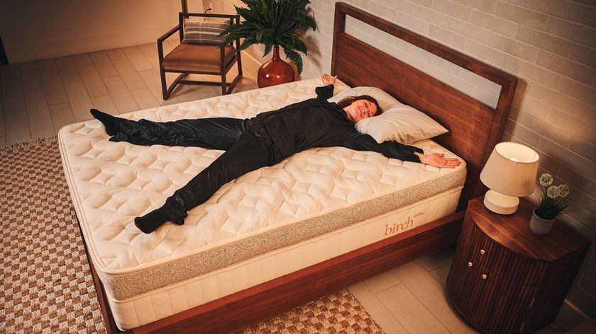 Our mattress tester wearing black silk pyjamas laying on her back on the Birch Elite mattress on wooden bedframe in warm lit bedroom with bedside plant, chair, unit, lamp and checkred rug